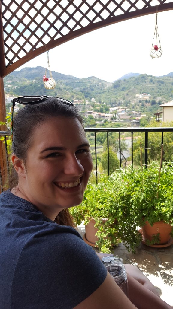Image of Briana turning back toward the camera and smiling. Her hair is pulled back with sunglasses on her head, and she is wearing a blue t-shirt. She is seated outside with a gazebo arch framing her and plants potted on the ground next to a wrought-iron fence. Beyond the fence and arch are buildings dotted along the mountain backdrop. 